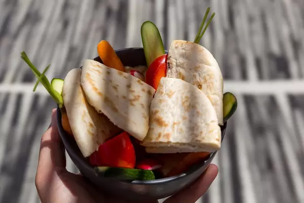 Hand holding a bowl with fresh cut veggies and pita for hummus in the shade