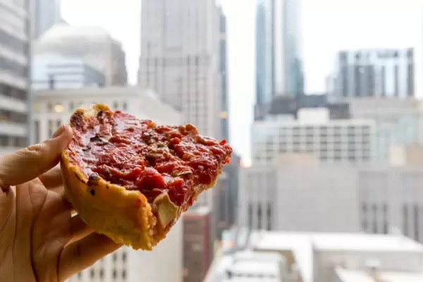 Hand holding a slice of Chicago-style pizza with cheese and tomato made by Pizzeria Uno, creators of the first deep dish pizza in 1943. In the background, the skyscrapers of Downtown Chicago