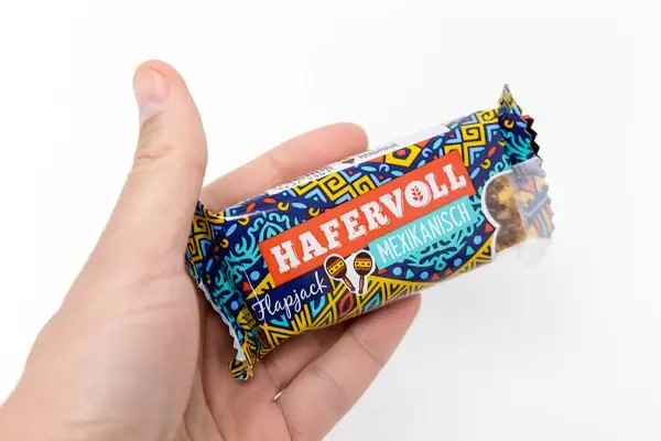 Hand holding "Hafervoll Flapjack Mexican" - cereal bar with colorful packaging showing two maracas in front of a white background