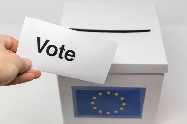 Hand holds a ballot paper with the text "Vote" in front of a white box with a European flag on it, as ballot box