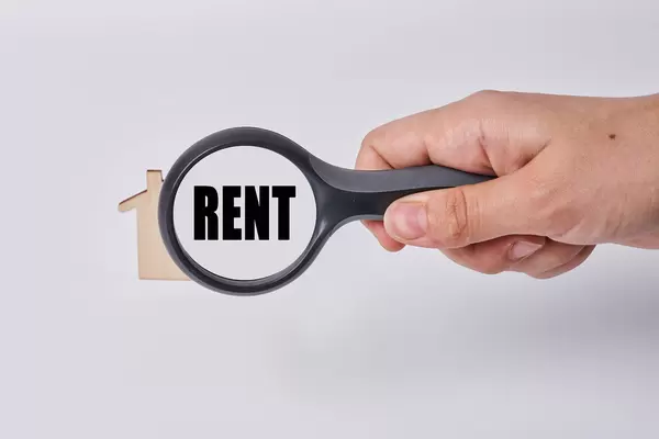 Hand holds magnifying glass over wooden house with text - rent. Concept of searching house for rent