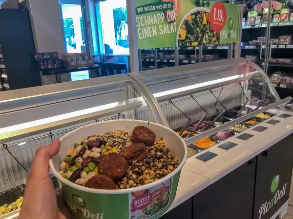 Hand holds To-Go-Box from PicaDeli salad bar, filled with power corn mix with pasta & vegetarian tomato-basil veggie-balls, in front of salat bar