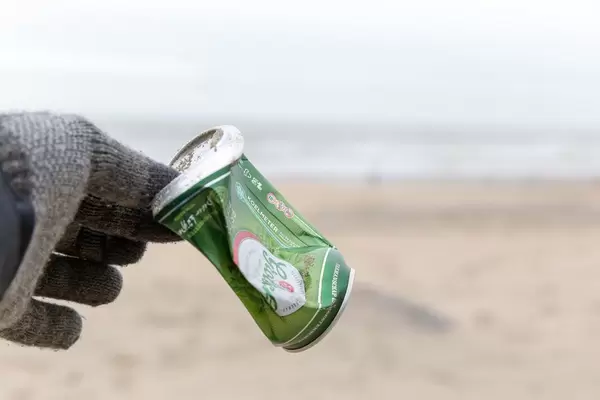 Hand in Handschuh hebt achtlos weggeworfene Bierdose vom Sandstrand auf