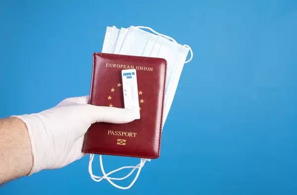 Hand in protective gloves holding passport with face masks and Covid-19 antigen test on blue background