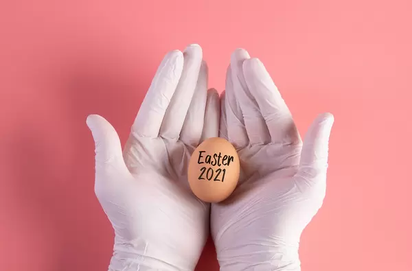 Hand in white protective gloves holding egg with Easter 2021 text