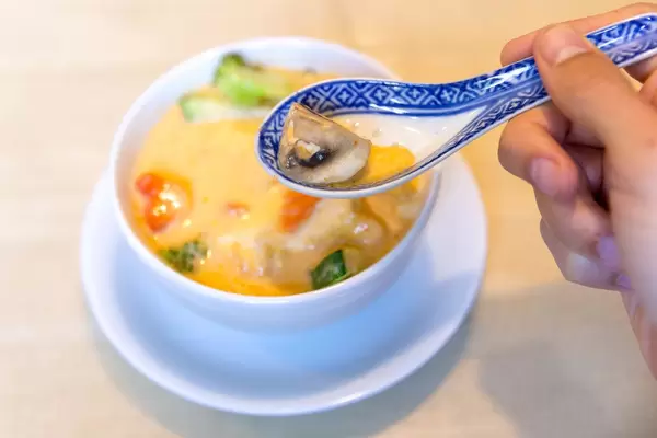 Hand mit einem asiatischen Porzellan-Löffel vor dem veganen Gericht "Tom Yum" Kokosmilchsuppe, mit Champions, Möhren, Zucchini und Brokkoli