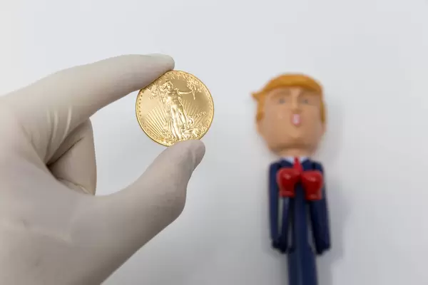 Hand mit Einmalhandschuh hält eine Unze Gold vor Trump-Figur mit Boxhandschuhen in der Coronakrise