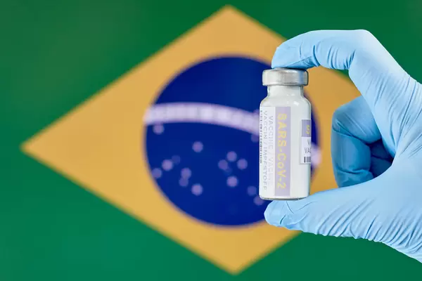 Hand mit Einmalhandschuhen hält Corona Impfstoff-Fläschchen vor der Flagge Brasiliens