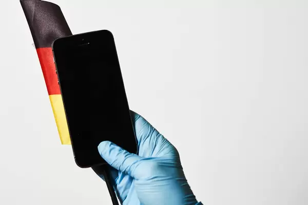 Hand mit Einweghandschuh hält Smartphone vor deutscher Flagge auf weißem Hintergrund mit Platz für Text