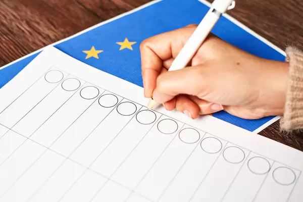 Hand mit Kugelschreiber vor einem leeren Stimmzettel mit der europäischen Flagge auf dem Tisch