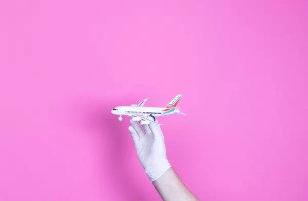 Hand mit medizinischen Handschuhen hält ein kleines Flugzeug vor pinkem Hintergrund