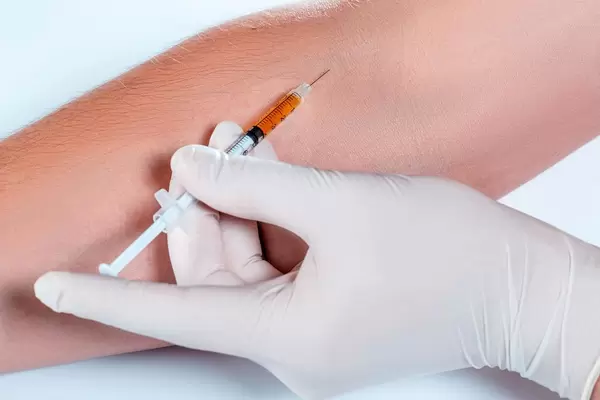 Hand mit weißen Handschuhen setzt Spritze in Armvene vor weißem Hintergrund