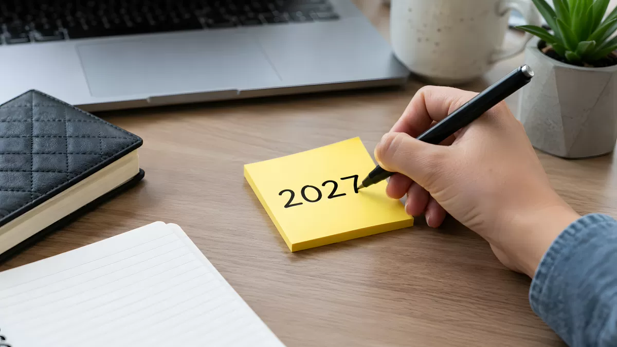 Hand schreibt 2027 auf gelben Notizzettel am Schreibtisch