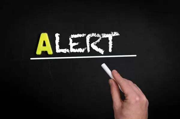 Hand schreibt das Wort "Alert" - warnen - mit Kreide auf eine schwarze Tafel