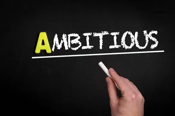 Hand schreibt das Wort "Ambitious" - Ambitioniert - mit Kreide auf eine schwarze Tafel