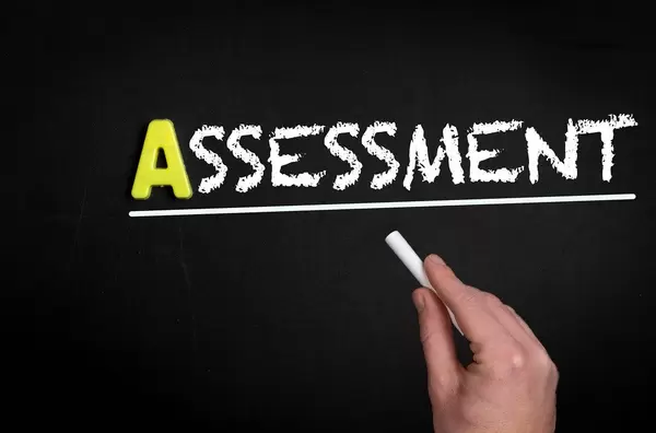Hand schreibt das Wort "Assessment" - Bewertung - mit Kreide auf eine schwarze Tafel