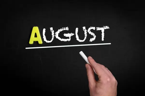 Hand schreibt das Wort "August" mit Kreide auf eine schwarze Tafel