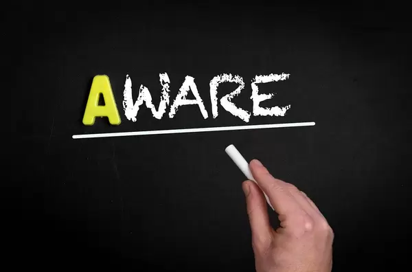Hand schreibt das Wort "Aware" - Bewusst - mit Kreide auf eine schwarze Tafel