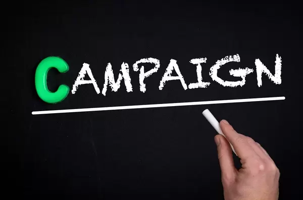 Hand schreibt das Wort "Campaign" - Kampagne - mit Kreide auf eine schwarze Tafel