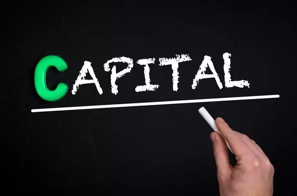 Hand schreibt das Wort "Capital" - Kapital - mit Kreide auf eine schwarze Tafel