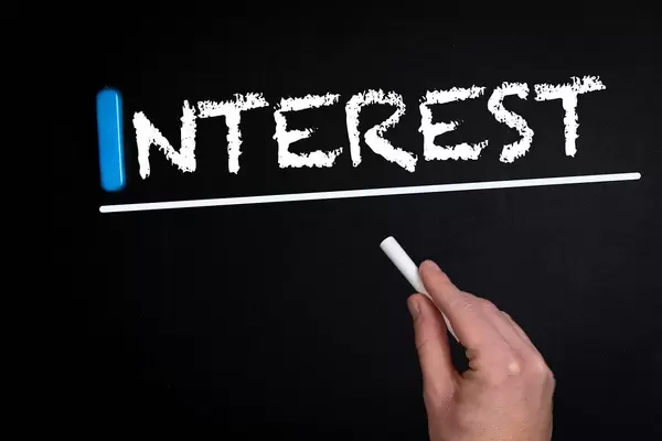 Hand schreibt das Wort "Interest" - Interesse - mit Kreide auf eine schwarze Tafel