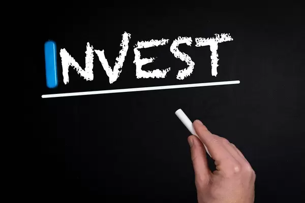 Hand schreibt das Wort "Invest" - investieren - mit Kreide auf eine schwarze Tafel
