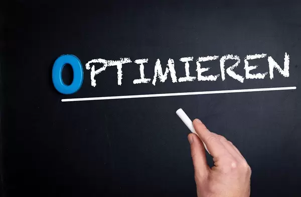 Hand schreibt das Wort "Optimieren" mit Kreide auf eine schwarze Tafel