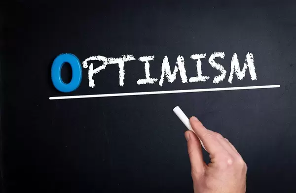 Hand schreibt das Wort "Optimism" mit Kreide auf eine schwarze Tafel