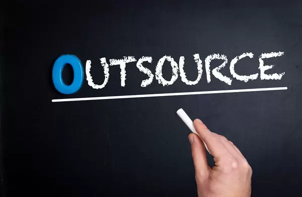Hand schreibt das Wort "Outsource" - Auslagern - mit Kreide auf eine schwarze Tafel
