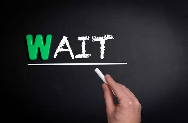 Hand schreibt das Wort "Wait" - Warte - mit Kreide auf eine schwarze Tafel