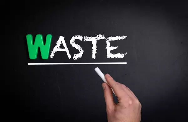 Hand schreibt das Wort "Waste" - Verschwendung - mit Kreide auf eine schwarze Tafel
