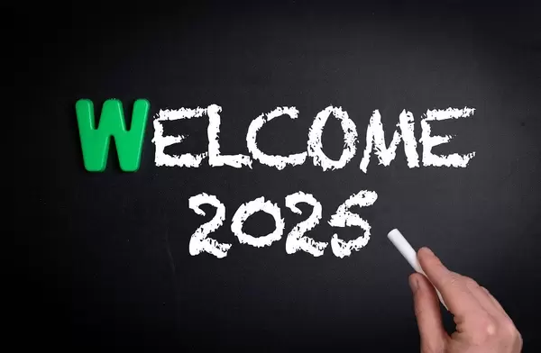 Hand schreibt das Wort "Welcome 2025" - Willkommen 2025 - mit Kreide auf eine schwarze Tafel