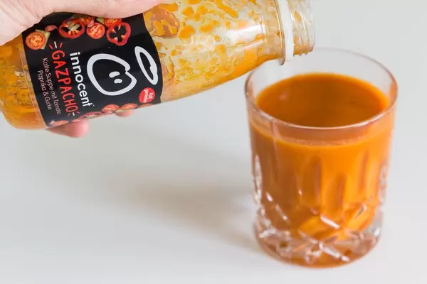 Hand schüttet die kalte Gemüsesuppe mit dem spanischen Namen "Gazpacho" von Innocent aus der kleinen Flasche in ein Glas vor weißem Hintergrund