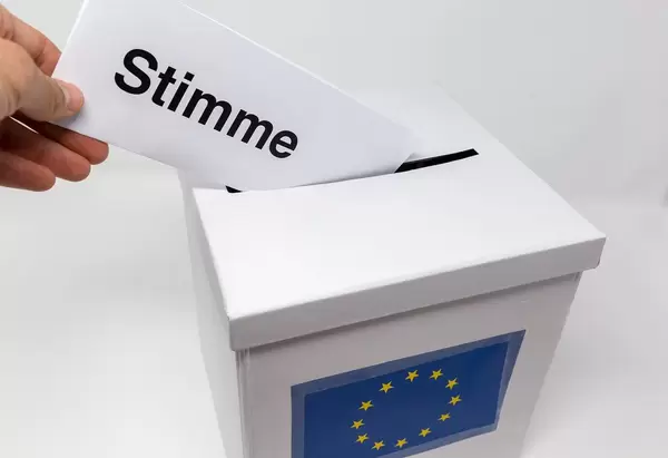 Hand steckt einen Wahlschein mit der Aufschrift "Stimme" in einen weißen Karton mit Europaflagge als Wahlurne