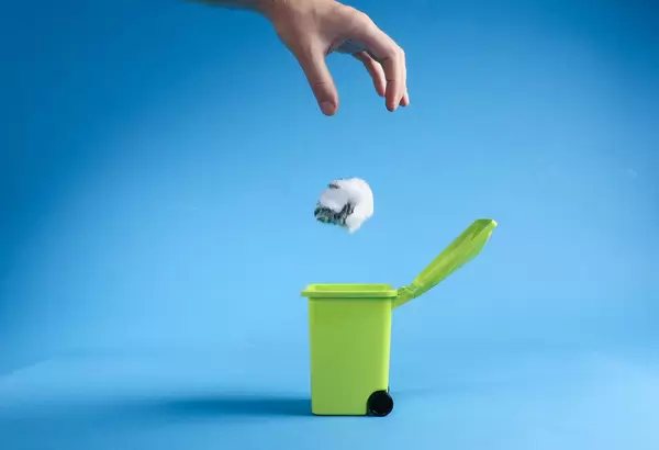 Hand throwing plastic bag into trash can on blue background