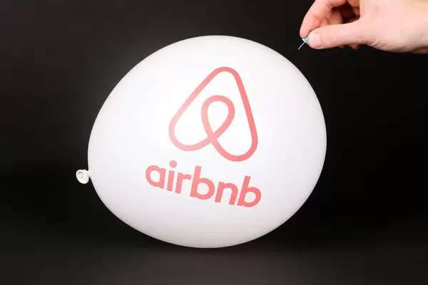 Hand uses a needle to burst a balloon with Airbnb logo