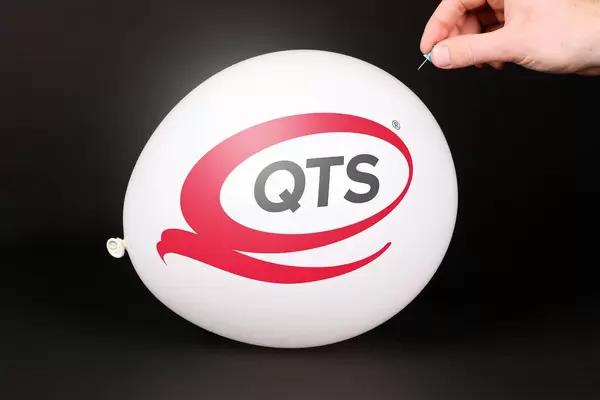 Hand uses a needle to burst a balloon with Quality Technology Services logo