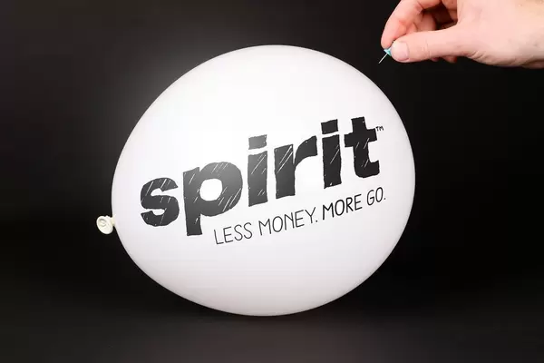 Hand uses a needle to burst a balloon with Spirit Airlines logo