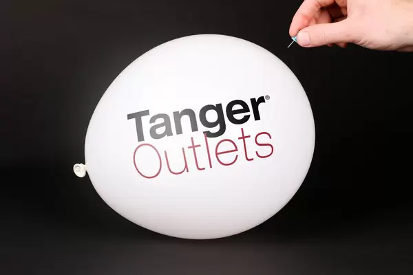 Hand uses a needle to burst a balloon with Tanger Outlets logo