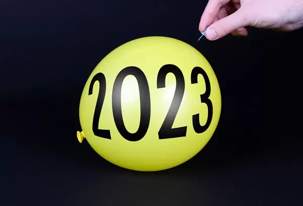 Hand uses a needle to burst a yellow balloon with 2023 text