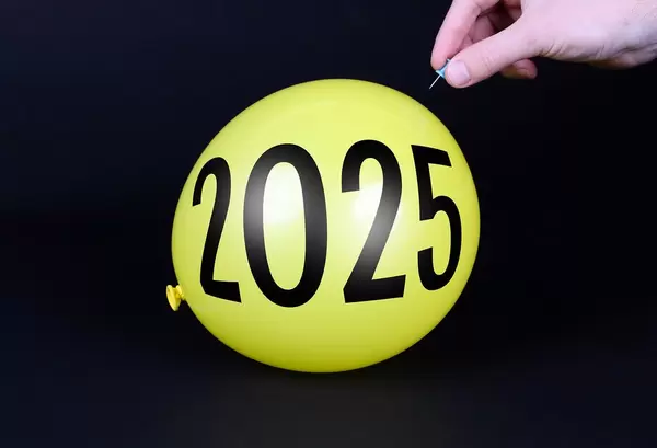 Hand uses a needle to burst a yellow balloon with 2025 text