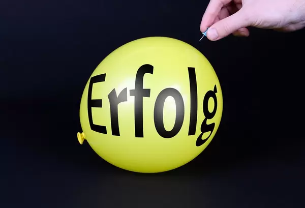Hand uses a needle to burst a yellow balloon with Erfolg text