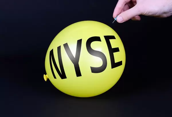 Hand uses a needle to burst a yellow balloon with NYSE text