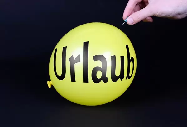 Hand uses a needle to burst a yellow balloon with Urlaub text