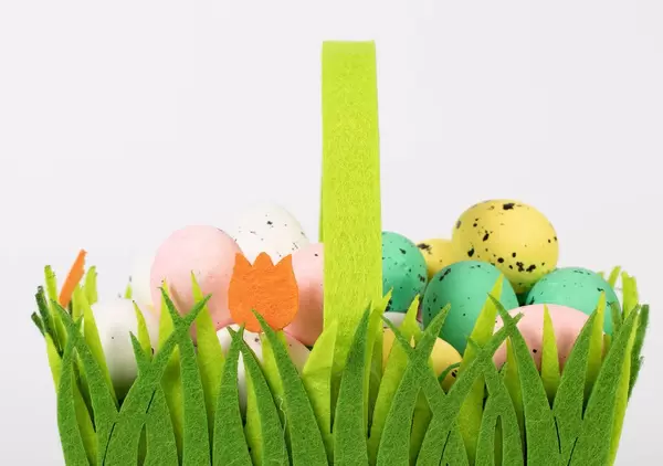 Handbemalte Ostereier in grünem, mit Gras und Tulpen verziertem Osterkorb vor weißem Hintergrund