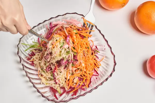 Hände mit Besteck mischen einen bunten Salat in einer quadratischen Glasschüssel