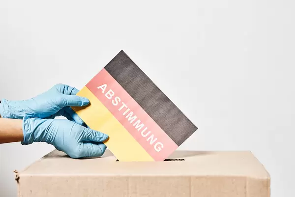Hände mit Einweghandschuhen werfen deutsche Flagge und Schrift "Abstimmung" in die Wahlurne ein