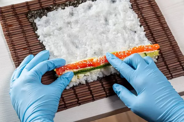 Hands in blue gloves spread the ingredients on a leaf of seaweed nori. Preparing sushi