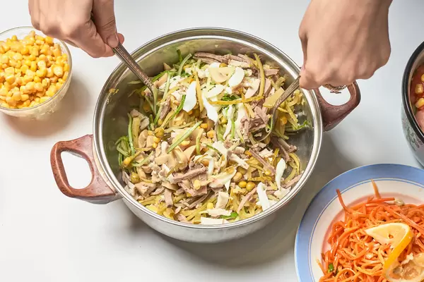 Hands using cutlery to mix the ingredients in a pot: mais, cucumber and meat cut in strips