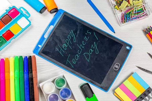 Happy Teacher's Day inscription with markers, pencils, paper clips, paints and compasses on a white wooden table. Top view